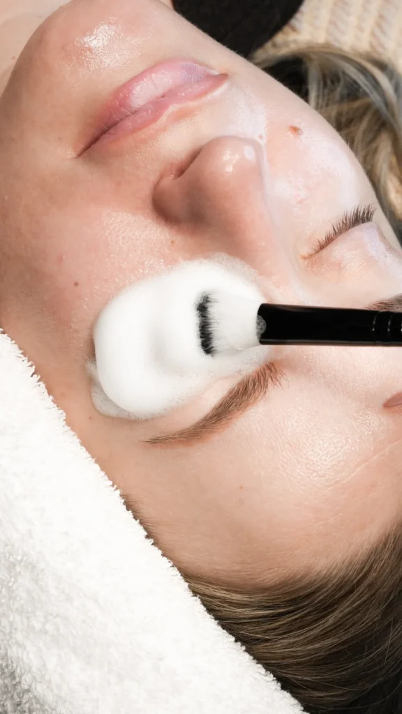 A person lying down with eyes closed while a soft brush applies cleansing foam on the eyelid area, demonstrating proper lash extension aftercare cleansing.